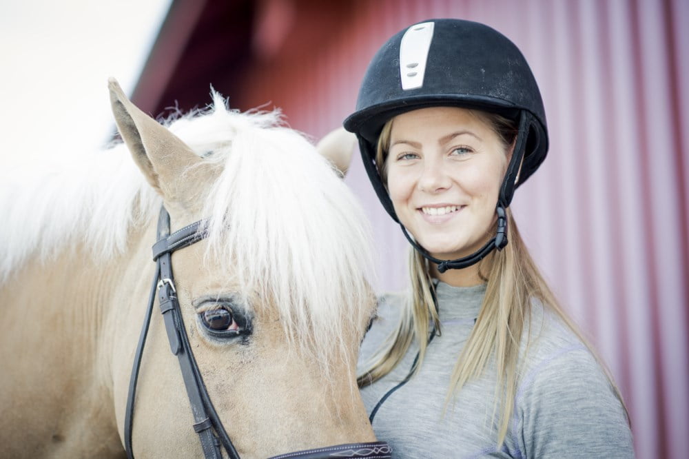 Smilende kvinne med hjelm står ved siden av en hest med hvit manke.