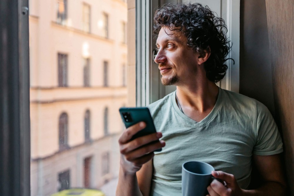Mann med mobil og kaffekopp som ser ut av vinduet