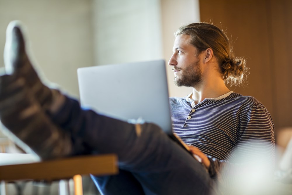 En ung mann sitter avslappet med føttene på et bord mens han bruker en laptop. Han har langt hår samlet i en liten hestehale, og han er kledd i en stripet genser.