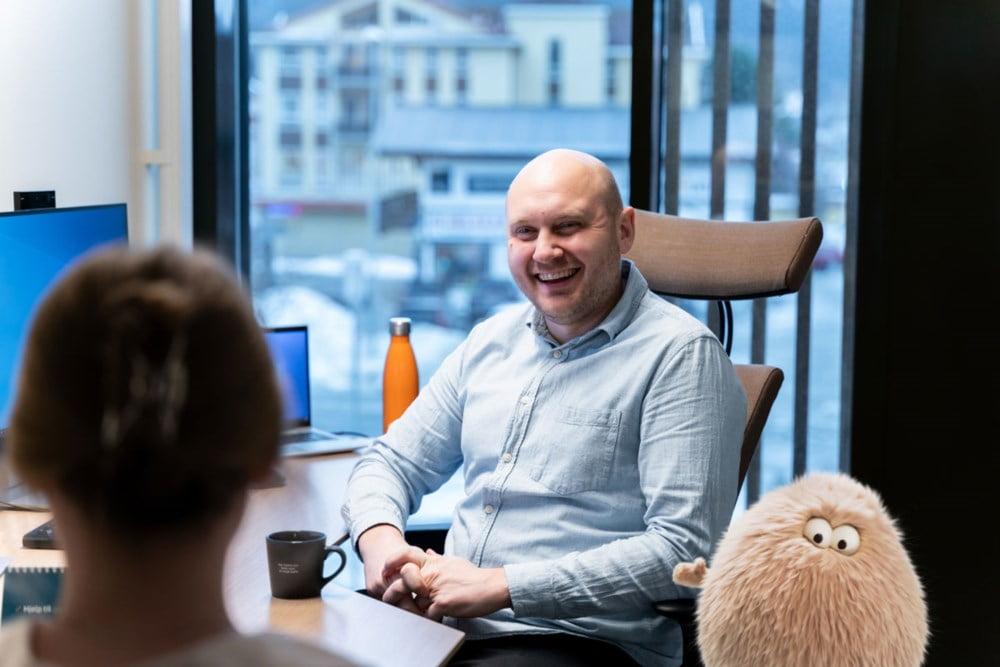 Mann sitter bak en pult og smiler til en kunde. En søt hårdott nede i hjørnet vinker til kamera.