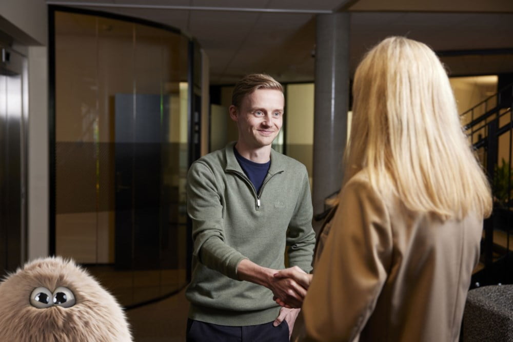 Ung, smilende mann håndhilser på en dame. I hjørnet ned til venstre titter en søt hårdott fram.
